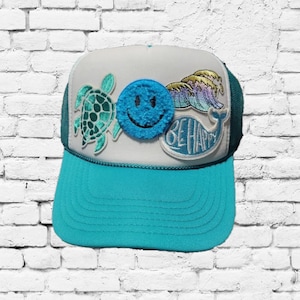 Può includere: Un cappello da camionista bianco e turchese con una varietà di toppe colorate. Le toppe includono un viso sorridente, una tartaruga marina, una balena e un'onda. Il testo "Be Happy" è sulla toppa della balena.