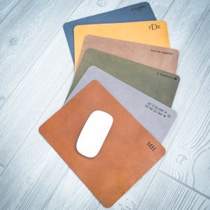 May include: A stack of six leather mousepads in various colors, each with a different personalized inscription. The top mousepad is dark blue with the initials "FDR" in white. The second mousepad is yellow with the text "Love thy neighbor" in black. The third mousepad is brown with the initials "A. Sanderson" in black. The fourth mousepad is green with the coordinates "41°2' 31.056" N 73°54' 21.744" W" in black. The fifth mousepad is gray with a heart symbol. The bottom mousepad is brown with the initials "MH" in black. A white computer mouse is on top of the bottom mousepad.