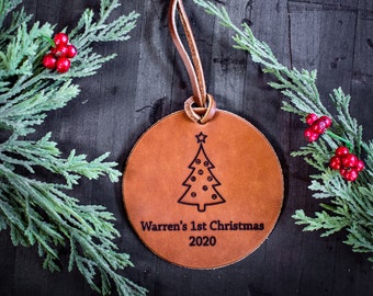 May include: A brown leather Christmas ornament with a star and a Christmas tree design. The ornament is personalized with the text "Warren's 1st Christmas 2020".
