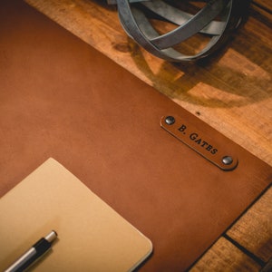 May include: A brown leather desk mat with a personalized nameplate that reads "B. GATES". The mat is on a wooden surface and has a notepad and pen on top.