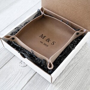 以下が含まれることがあります： 正面に「M&S」のイニシャルと「est. 2019」の刻印が施された茶色の革製の小物入れ。白い箱の中に黒い破り紙と一緒に収められています。