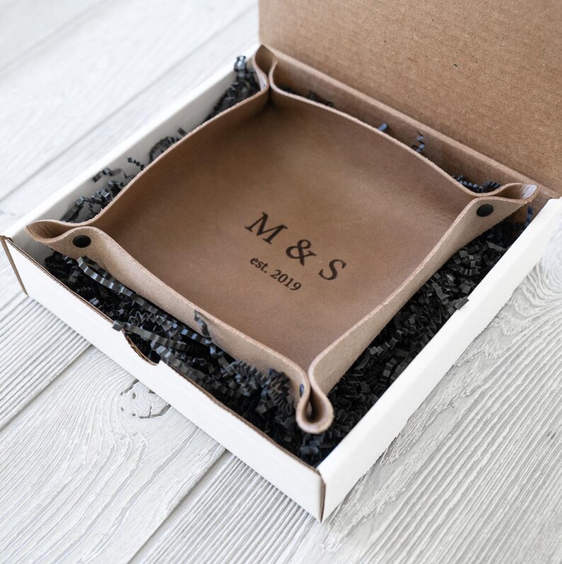 以下が含まれることがあります： 正面に「M&S」のイニシャルと「est. 2019」の刻印が施された茶色の革製の小物入れ。白い箱の中に黒い破り紙と一緒に収められています。