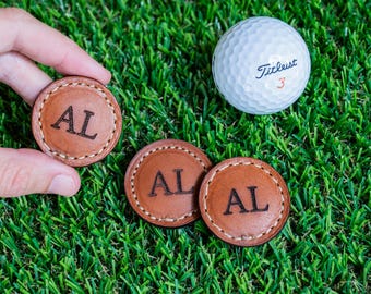 May include: Three brown leather golf ball markers with the initials "AL" engraved on each. The markers are on a green artificial grass surface. A white golf ball with the text "Titleist 3" is in the background.