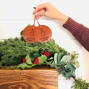 May include: Two brown leather Christmas ornaments with a tree design and personalized text. The ornament on the left says "The Millers Christmas '19" and the ornament on the right says "The Leonards Est. 2009".