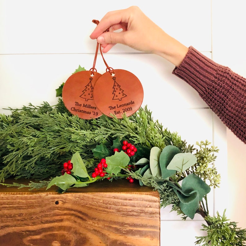 May include: Two brown leather Christmas ornaments with a tree design and personalized text. The ornament on the left says "The Millers Christmas '19" and the ornament on the right says "The Leonards Est. 2009".