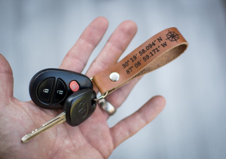 May include: A brown leather keychain with a compass design and the coordinates "30&deg; 19' 58.094' N 87&deg; 08' 59.171' W" engraved on it. The keychain is attached to a black car key.