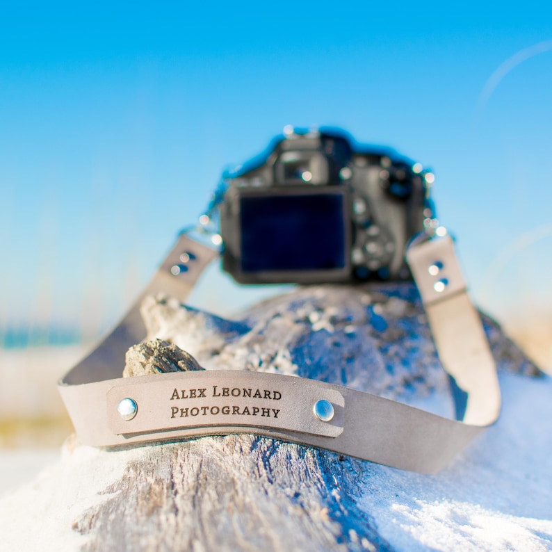 May include: A light gray leather camera strap with the text "ALEX LEONARD PHOTOGRAPHY" engraved on it. The strap is attached to a black camera lens.