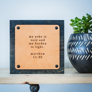 Puede incluir: Un pequeño letrero de madera con una placa de cuero marrón. La placa tiene el texto "my yoke is easy and my burden is light. matthew 11:30" impreso en ella.