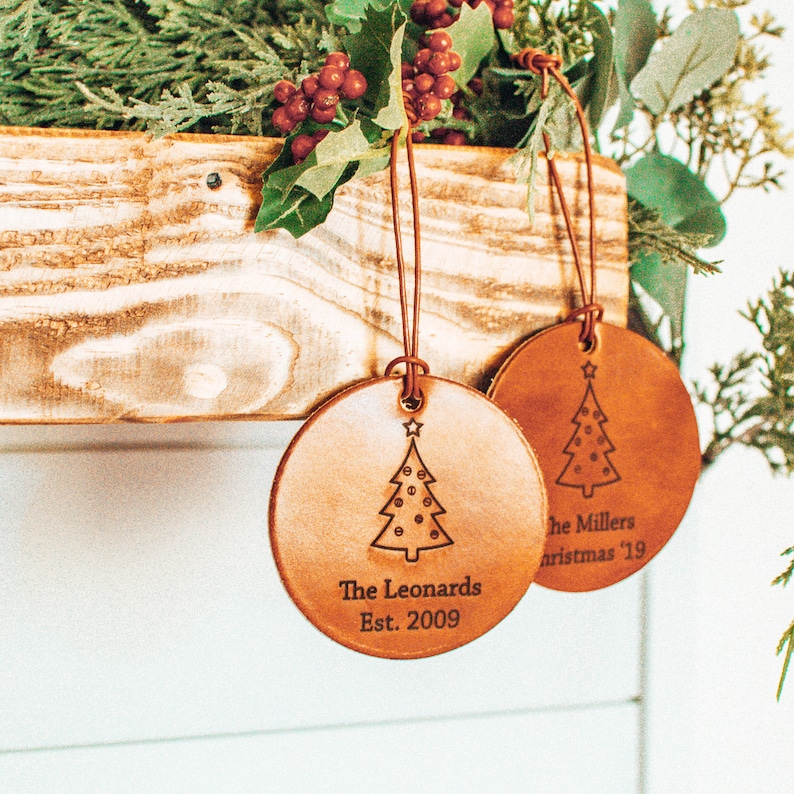 May include: Two brown leather Christmas ornaments with a tree design and personalized text. The first ornament reads "The Leonards Est. 2009". The second ornament reads "The Millers Christmas '19".