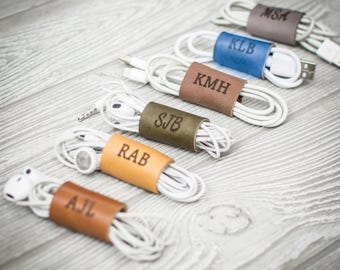 May include: Six white wired earbuds and charging cables, each neatly coiled and secured with a personalized leather cord organizer. The organizers are in various colors including tan, light brown, olive, dark brown, blue, and gray, each embossed with three-letter initials: AJL, RAB, SJB, KMH, KLB, and MSA.