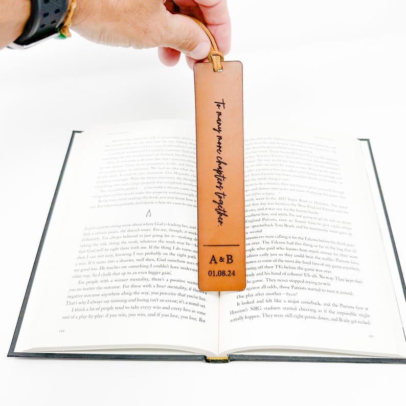 A hand holding a personalized leather bookmark with the text "To many more chapter together" engraved on it, resting on an open book.