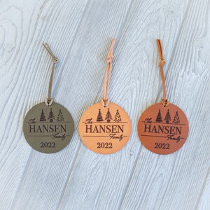 May include: Three leather Christmas ornaments with a brown leather cord. Each ornament is a circle with a design of three stylized Christmas trees and the text "The Hansen Family 2022". The ornaments are in three different colors: green, brown, and orange.