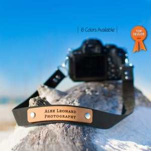 Op de afbeelding: Zwarte cameraband met een bruin leren patch waarop "Alex Leonard Photography" staat. De band ligt op een stuk drijfhout met een camera op de achtergrond. De tekst "6 kleuren beschikbaar" staat bovenaan in het midden van de afbeelding. Een "Top Seller" badge staat rechtsboven in de afbeelding.