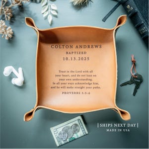 May include: A square tan leather valet tray with four snapped corners. The centre is engraved with the text: COLTON ANDREWS BAPTIZED 10.13.2025. Below this, the text reads: Trust in the Lord with all your heart, and do not lean on your own understanding. In all your ways acknowledge him, and he will make straight your paths. PROVERBS 3:5-6.