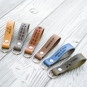 May include: Six leather keychains with different engravings. The keychains are brown, gray, blue, and green. The engravings include names, dates, coordinates, and a compass symbol.