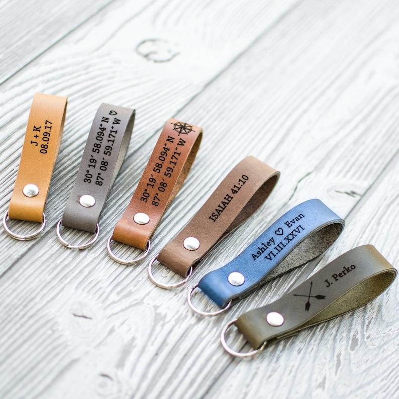May include: Six leather keychains with different engravings. The keychains are brown, gray, blue, and green. The engravings include names, dates, coordinates, and a compass symbol.
