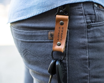 May include: A brown leather keychain with a metal ring and a black metal clip. The keychain has the coordinates "35° 58' 38.405" N 92° 17' 54.952" W" engraved on it.