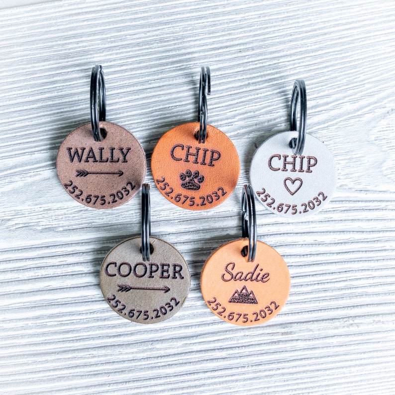Puede incluir: Cinco chapas redondas de cuero para perros con anillos de metal. Las chapas son de color marr&oacute;n, naranja y blanco con diferentes dise&ntilde;os y texto. El texto en las chapas incluye los nombres "Wally", "Chip", "Chip", "Cooper" y "Sadie" junto con n&uacute;meros de tel&eacute;fono.