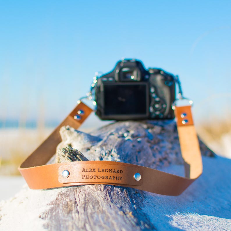 May include: A brown leather camera strap with the text "ALEX LEONARD PHOTOGRAPHY" embossed on it. The strap is attached to a black camera lens.