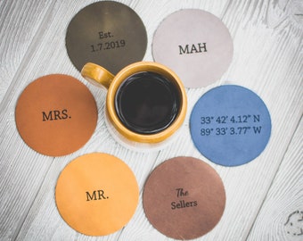 May include: A yellow mug filled with coffee is surrounded by six personalized leather coasters. The coasters are in various colors, with text including "Est. 1.7.2019", "MAH", "MRS.", "33° 42' 4.12" N 89° 33' 3.77" W", "MR.", and "The Sellers".