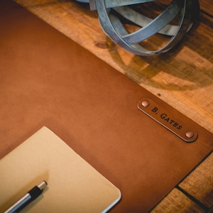 May include: A brown leather desk mat with a personalized name tag that reads "B. GATES". A notepad and pen are on the mat.