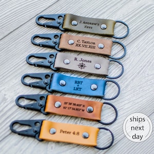 May include: Six leather keychains with black metal lobster clasps and rings. Each strap features two silver rivets and custom engravings: J. ANDREW'S KEYS, C. TAYLOR XX.VII.XIII, R. Jones with a compass, RBT+LKT, coordinates, and Peter 4:8.