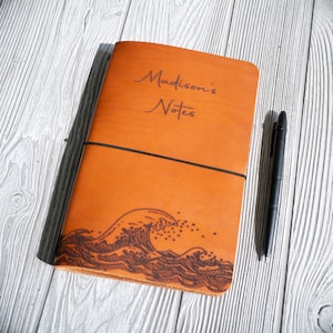 May include: A brown leather journal with an elastic closure and a wave design on the front. The journal is labeled "Madison's Notes".