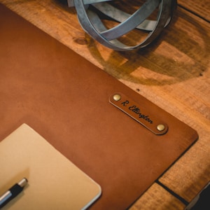May include: A brown leather desk mat with a personalized nameplate that reads "R. Ellington". The mat is on a wooden surface.