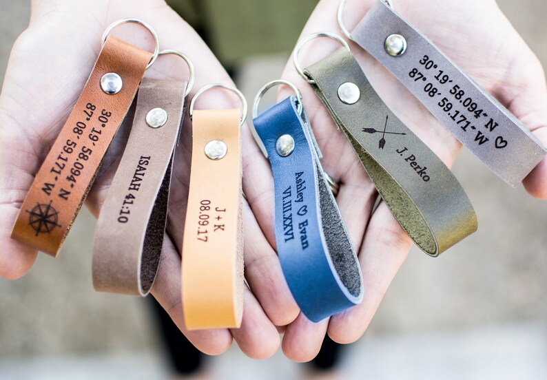 May include: Six leather keychains with different engravings. The keychains are brown, tan, orange, blue, and gray. The engravings include names, dates, coordinates, and a compass.