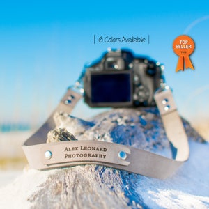 May include: A light gray leather camera strap with a personalized nameplate that reads "ALEX LEONHARD PHOTOGRAPHY". The strap is attached to a black camera lens and is resting on a piece of driftwood. The background is a bright blue sky with white clouds.