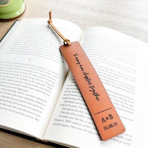 An open book with a leather bookmark featuring a personalized message, resting on a wooden surface.