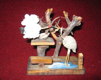 May include: A wooden sculpture of two white cranes perched on a tree branch. The cranes are made of white beads and the tree is made of natural branches. The sculpture is on a wooden base with a painted blue pond and a small wooden sign that says "石山".