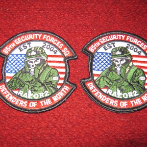 May include: Two black and red embroidered patches with a soldier in a green camouflage uniform holding a rifle. The patches are circular and have the text "85th Security Forces SQ EST 2004 DEFENDERS OF THE NORTH RAZOR2" around the edge.