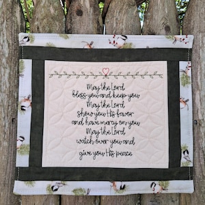 May include: A quilted wall hanging with a cream-colored center featuring embroidered text: "May the Lord bless you and keep you..." The border is a light green fabric with bird and berry patterns, framed by a darker green border. A red heart is embroidered above the text.