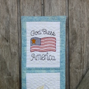 May include: A blue and white quilted wall hanging with three panels. The top panel features a red, white, and blue American flag with the text "God Bless America". The middle panel features a sailboat with a yellow sail. The bottom panel features a slice of watermelon with the text "Sweet Sweet Summertime".