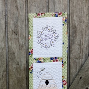 May include: A quilted wall hanging with three embroidered panels. The top panel says "Hello Spring" within a floral wreath. The middle panel features a beehive with a bee. The bottom panel shows a watering can with flowers and the word "Blessings".