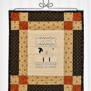 May include: A beige and brown quilted wall hanging with a black frame. The center of the hanging features a white sheep with black legs and a black head. The text "Live Simply" is embroidered in black thread above the sheep. The alphabet is embroidered in black thread below the sheep.