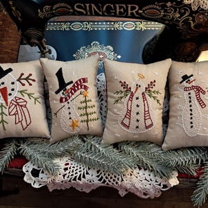 May include: Four rectangular decorative pillows with embroidered snowman designs. Each pillow features a different snowman with a black hat, scarf, and other festive details. The pillows are on a white lace doily with pine branches.
