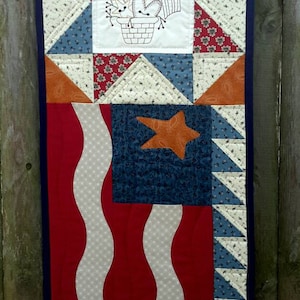 May include: A red, white, and blue patriotic quilt with a star and a basket of flowers embroidered in the center. The quilt is made of patchwork fabric and has a wavy design.