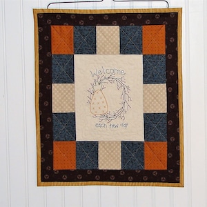 May include: A quilted wall hanging with a welcoming message. The central panel features an embroidered pear-shaped design within a wreath, with the words "Welcome each new day." The quilt has a patchwork design in shades of blue, orange, and cream, framed by a dark brown border.