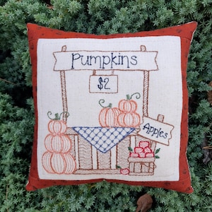 May include: Embroidered pillow featuring a fall harvest scene. The design includes a sign that reads "Pumpkins" and "Apples", with a price tag of $2. The artwork depicts stacks of pumpkins and a basket of apples, all in warm autumnal colors.