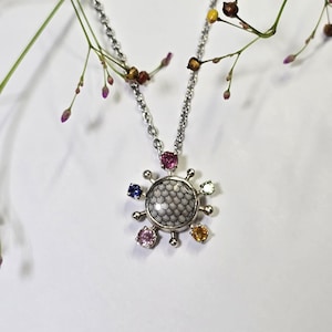 Puede incluir: Un collar de plata con un colgante circular que presenta un patrón de escamas blancas y negras en el centro. El colgante está rodeado por pequeñas piedras preciosas engastadas en tonos rosa, azul, amarillo y verde, unido a una fina cadena.