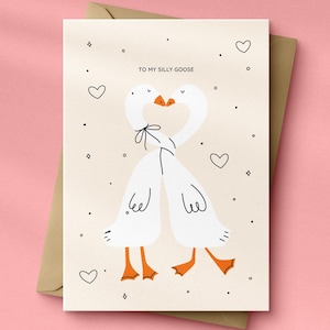 May include: Greeting card with a whimsical illustration of two white geese with orange feet and beaks, necks intertwined. The card reads "TO MY SILLY GOOSE" with hearts and dots on a beige background, set against a brown envelope on a pink surface.