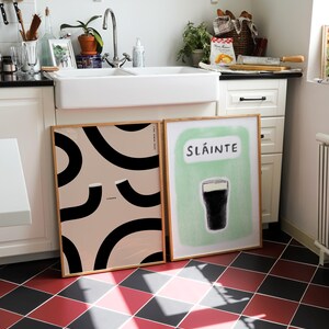 Slainte Guinness print hand drawn illustration of a guinness pint glass with text that says Sláinte hand drawn in charcoal and pastel oil. Sat next to our other abstract guinness print on a kitchen floor