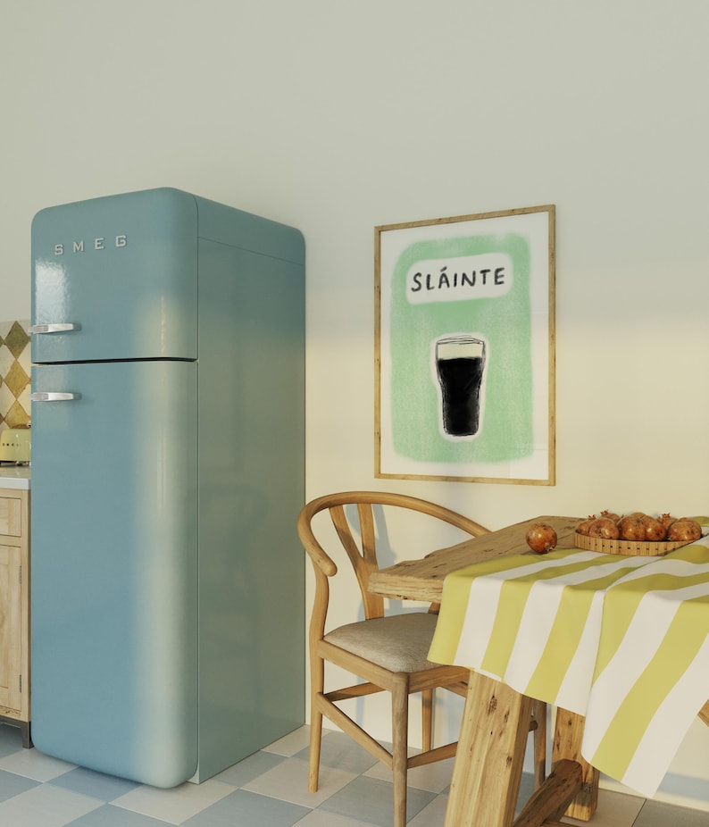 May include: A light blue retro refrigerator with the word "SMEG" on the front. A framed print with the word "Sl&aacute;inte" and a drawing of a glass of beer hangs on the wall. A wooden table with a yellow and white striped tablecloth sits in front of the refrigerator.