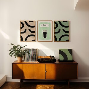 May include: Three framed prints on a white wall. The center print features a green background with the word "Sl&aacute;inte" and a black silhouette of a glass of beer. The prints on either side feature abstract black and white designs on a light beige and green background.