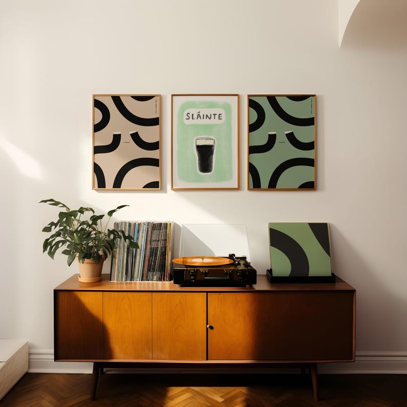 May include: Three framed prints on a white wall. The center print features a green background with the word "Sl&aacute;inte" and a black silhouette of a glass of beer. The prints on either side feature abstract black and white designs on a light beige and green background.