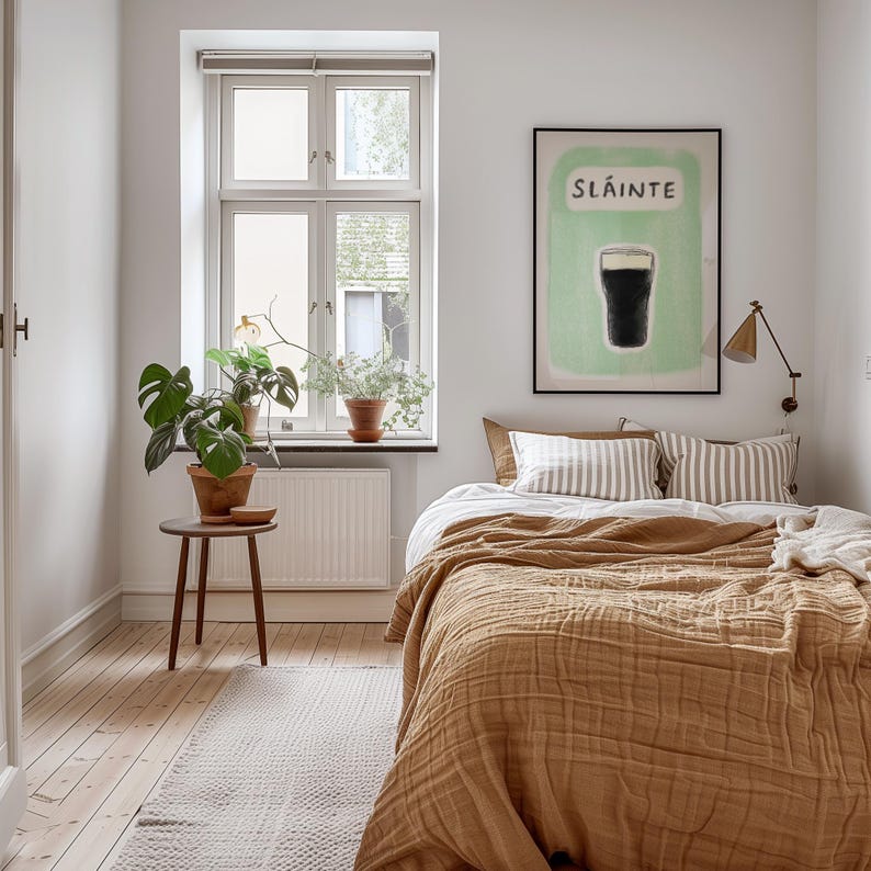 May include: A bedroom with a brown bed with a white and brown striped pillow and a brown blanket. There is a framed print on the wall with the word "Sl&aacute;inte" and a glass of dark liquid. There is a plant in a pot on the windowsill and a small wooden stool with a plant on it.
