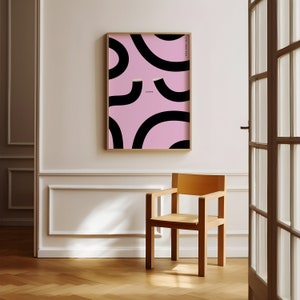 May include: A framed print with a pink background and black abstract shapes. The print is hanging on a white wall above a wooden chair.