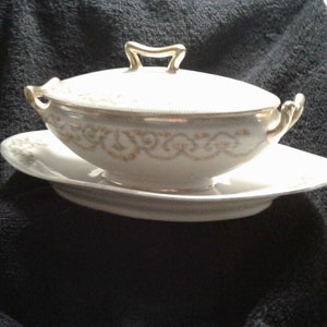 May include: White porcelain gravy boat with a gold trim and lid. The boat has a decorative pattern of gold swirls. The boat is sitting on a matching saucer.